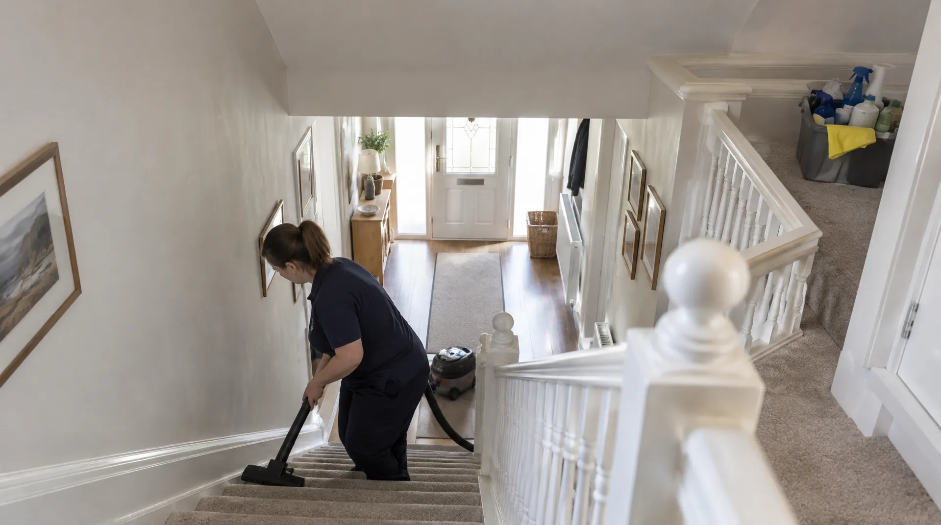 Cleaner vacuuming a carpeted staircase during a whole-house clean, with a supplies caddy on the landing